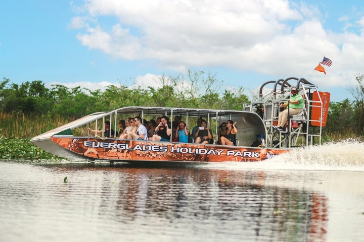 An airboat with passengers glides over water in a marshy area.