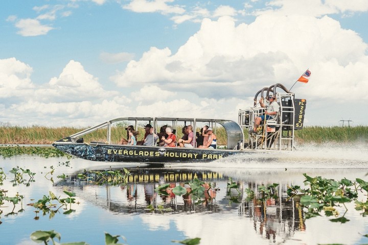Airboat with passengers glides on water in a marsh under a cloudy sky.