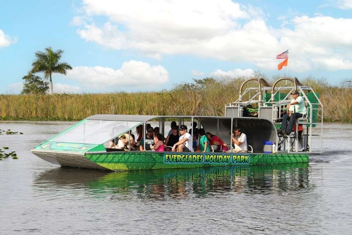 Airboat with passengers on a river surrounded by grass and palm trees, clear sky above.