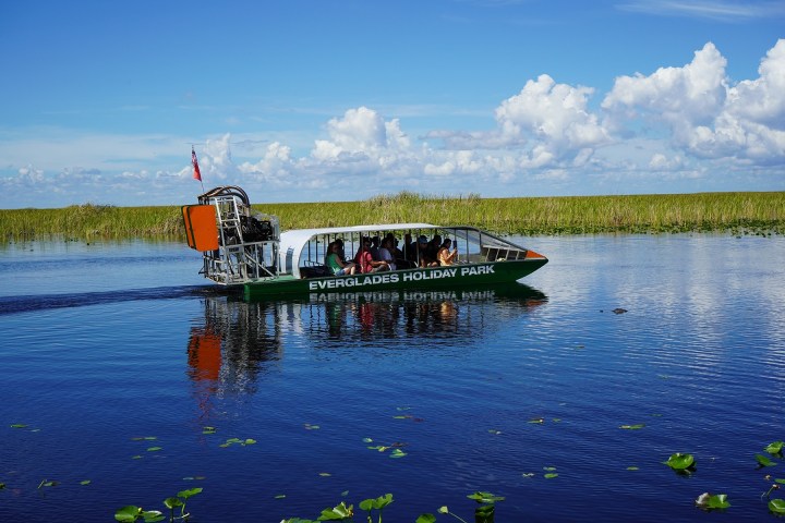 Airboat with tourists on a calm waterway in a grassy wetland area under a blue sky with clouds.